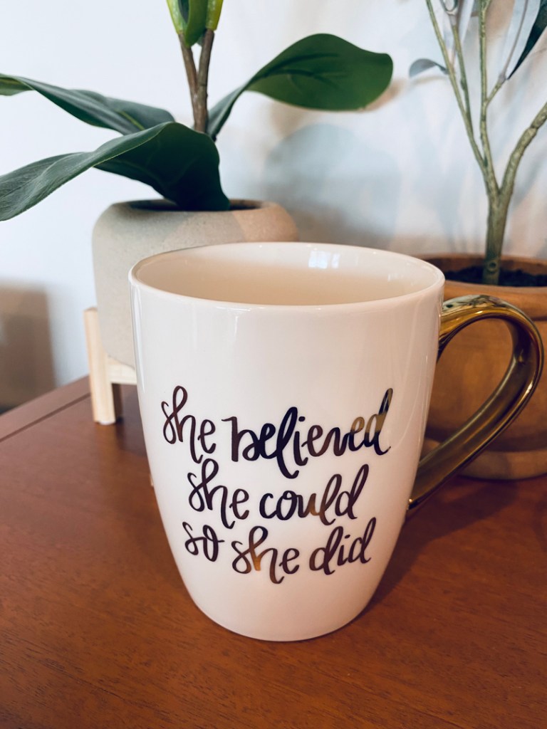 White mug with metallic gold-colored handle printed with gold script sitting on wooden table with two plants behind it. Inscription reads: "She believed she could so she did" 